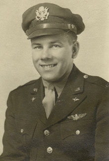 Black and white posed headshot of man wearing a uniform of a jacket, shirt, tie and hat. There are silver wings on the upper left-side of the jacket, and a WWII-era Air-Force silver officer insignia badge on the center of hat.  There is an aviation insignia pin and on the jacket lapel, and a single bar on the shoulder lapel. Subject is looking right in lens and is smiling