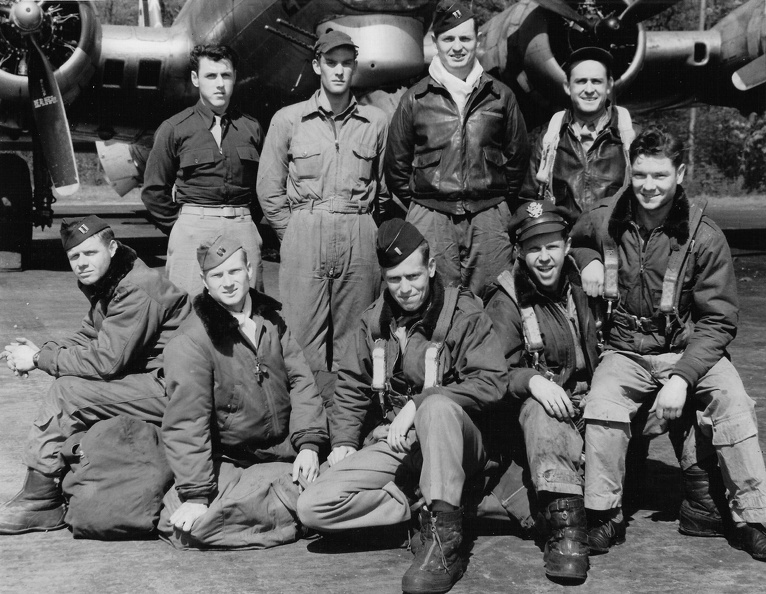 Nine men in Air Force uniform posed in two rows in front of a military airplane.  Five men standing to the rear and four men sitting in front on top of flying gear.  