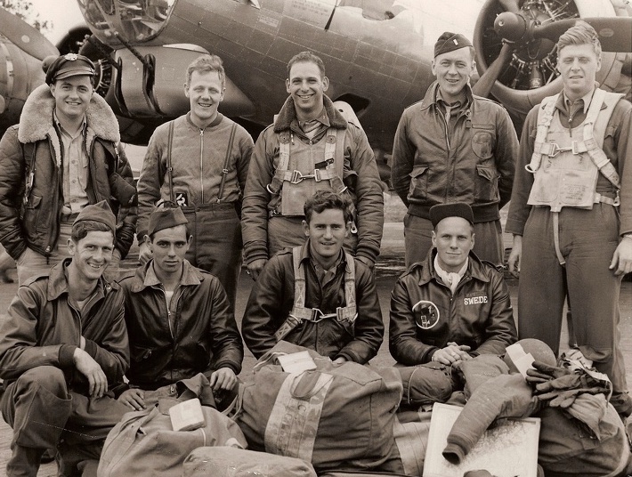 Nine men in Air Force uniform posed in two rows in front of a military airplane.  Five men standing to the rear and four men kneeling in front.  Men are wearing cold-weather flying gear.  There are canvas duffle bags on the ground in front of the men. 