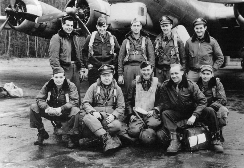 Ten men in Air Force uniform posed in two rows in front of a military airplane.  Five men standing to the rear and five men sitting in front on top of flying gear.  