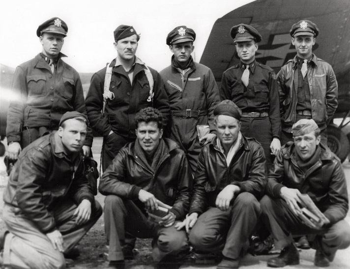 Nine men in Air Force uniform posed in two rows in front of a military airplane.  Five men standing to the rear and four men kneeling in front.  