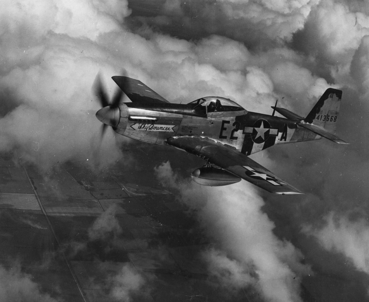 A P-51 Mustang (E2-A, serial number 44-13568) nicknamed "Sky Bouncer" of the 361st Fighter Group flies over the countryside. Image stamped multiple times on reverse: "Official Photograph US Army Air Forces", "For Personal Use Only" and "Passed by US Army Examiner, 10143." Handwritten caption on reverse: 'P-51 D, 361 FG (USAAF).'