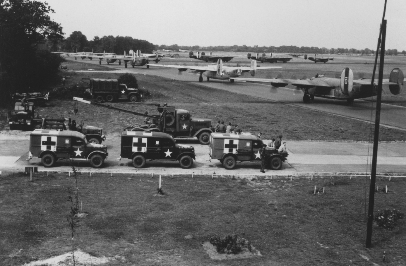 Field Ambulances standby as B-24 Liberators of the 44th Bomb Group line up for takeoff at Shipdham. Image has been signed on reverse with an illegible signature. Handwritten caption on reverse: '281-66-76.'