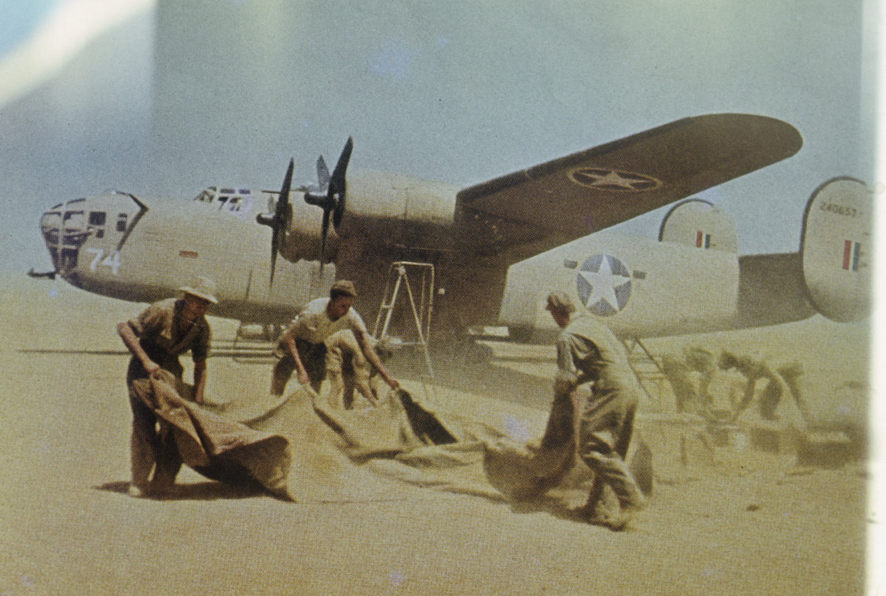 Personnel of the 376th Bomb Group lift dust sheets to protect their equipment infront of a B-24 Liberator (serial number 42-40657) nicknamed "GI Ginnie".