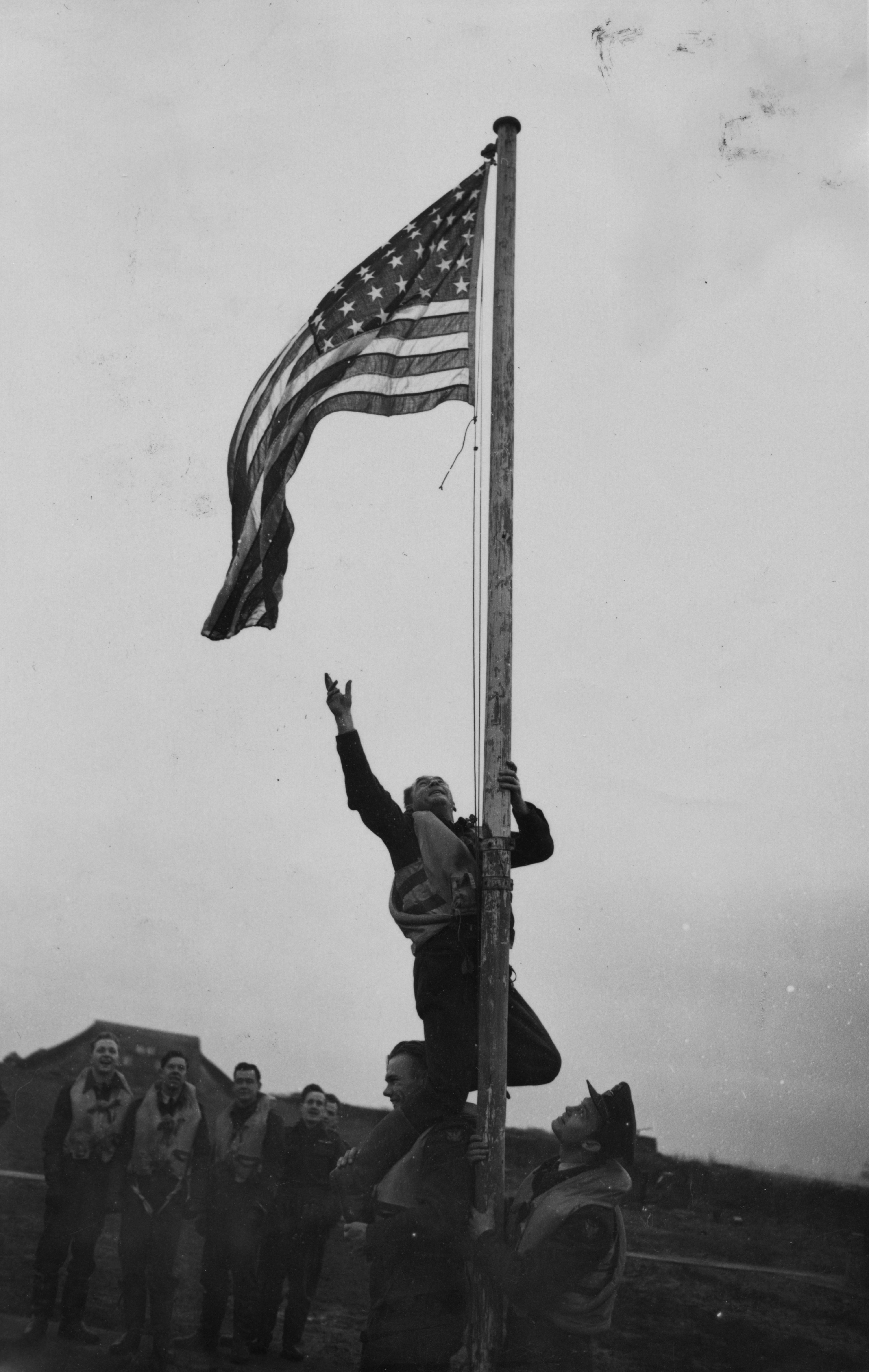 Pilot Officer Kenneth Le Roy Holder of No. 121 Eagle Squadron, RAF sits on the shoulders of Pilot Officer Donald Wilson MacLeod in order to reach the Stars and Stripes , 27 November 1941. Passed for publication 27 Nov 1941. Printed caption on reverse: 'Not For Publication Before 00.30 Hrs Sat. Nov 29th. First Pictures Of Second Eagle Squadron. 27.11.42. These are the first pictures to be taken of the second of the three American Eagle Squadrons to be formed in this country as units of the RAF Fighter Comm