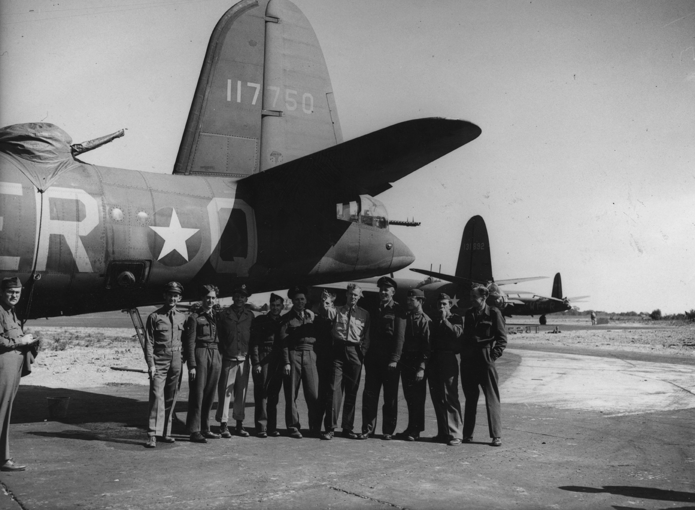 Personnel of the 322nd Bomb Group, including Flight Lieutenant Joe McCarty, talk with RAF personnel under the tail of a B-26 Marauder (serial number 41-17750) in August 1943. Passed for publication 18 Aug 1943. Printed caption on reverse: 'America's New Super Medium Bomber In Britain. 18.8.43. The U.S. Super Medium bomber, the "Marauder" is now operating from Britain after doing fine work in N. Africa and Sicily. It has a range of 2,000 miles, with a speed of 350 m.p.h. and a wing span of 65 ft., and carr