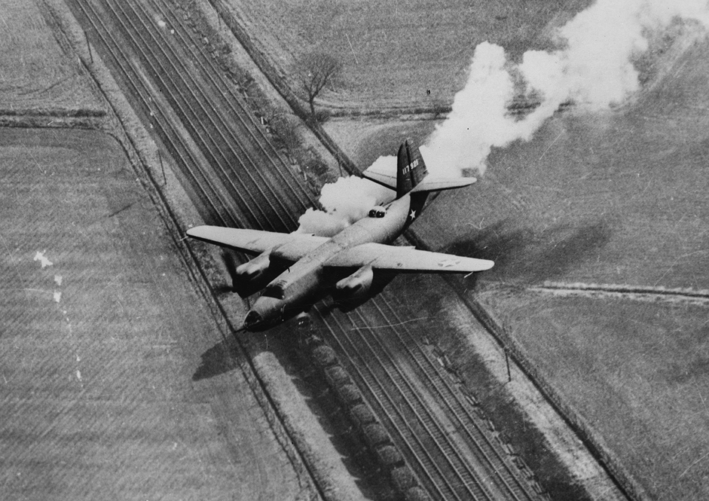 A B-26 Marauder (ER-C, serial number 41-17921) nicknamed "Mary V" of the 450th Bomb Squadron, 322nd Bomb Group flies low, trailing smoke, March 1943. Handwritten caption on reverse: '117921, ER:C.' Printed caption on reverse: "Appearances are deceiving. No, this Marauder is not on fire - the white smoke is actually coming from the locomotive on the tracks below. The picture was taken by an aerial photographer in another B-26 while the planes were flying over the English countryside, returning from a raid 