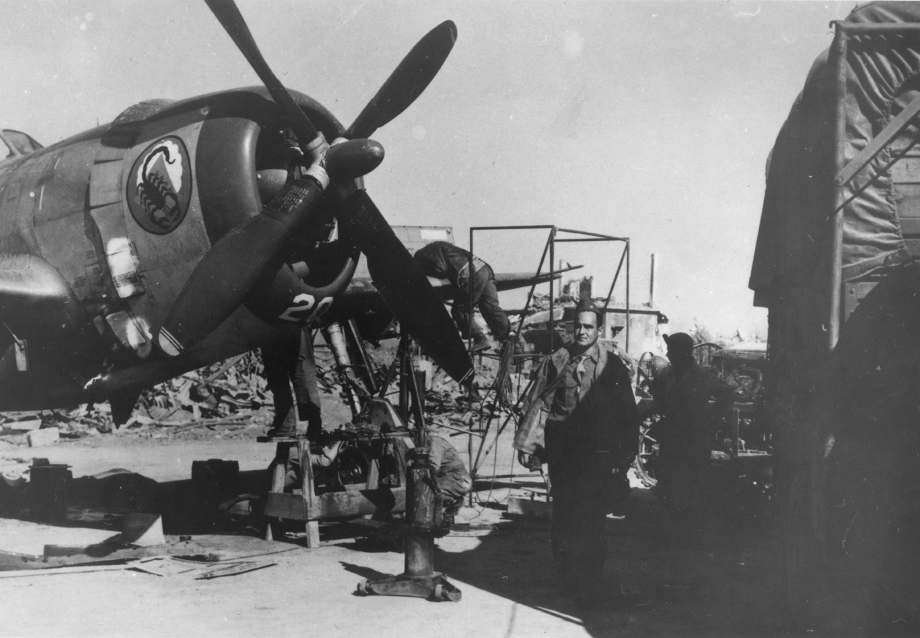 Ground personnel of the 57th Fighter Group work on the engines of a P-47 Thunderbolt, with the insignia of the 64th Fighter Squadron on its nose.