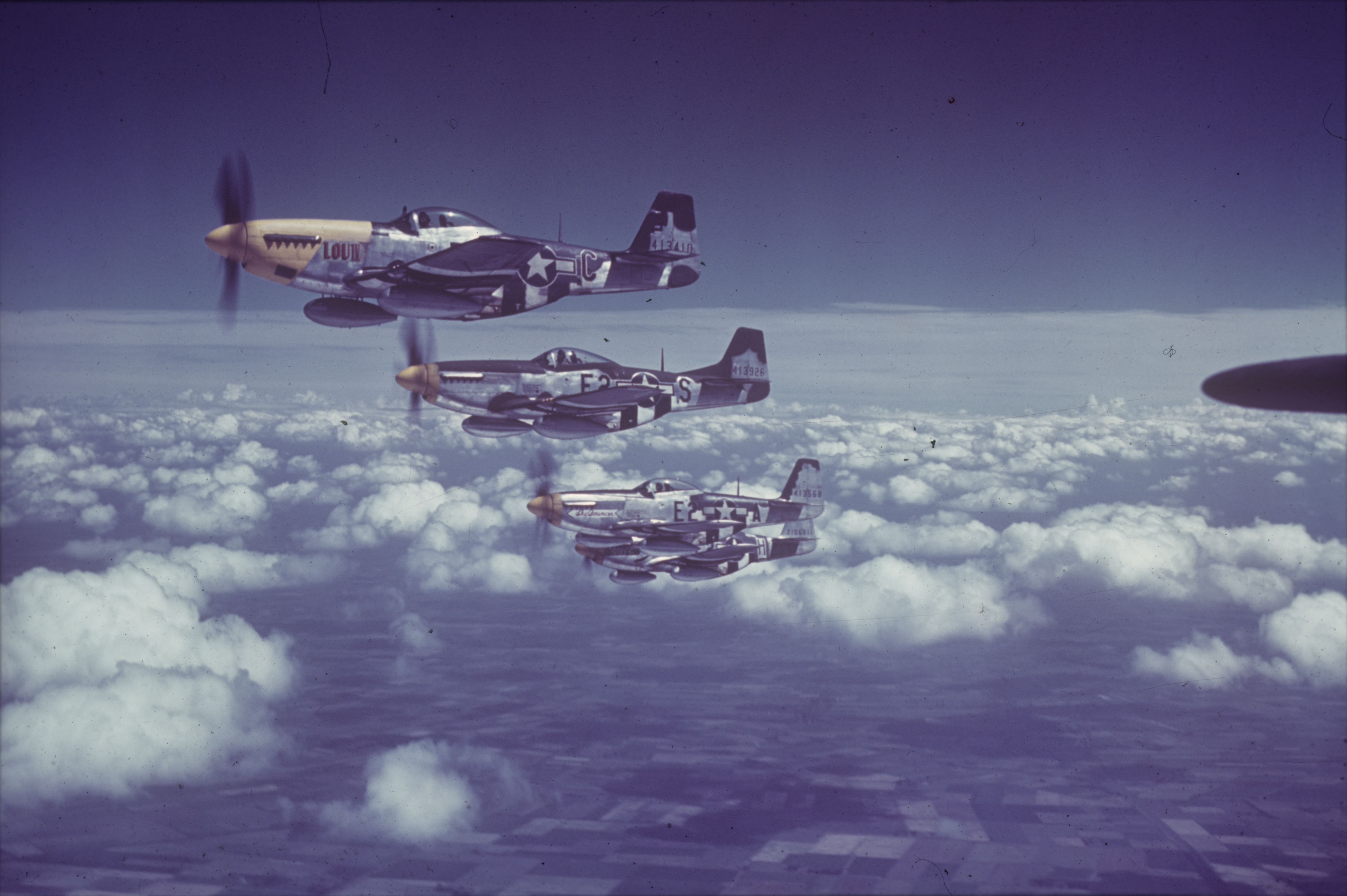 P-51 Mustangs (E2-C, serial number 44-13410) nicknamed "Lou IV (Athelene)", (E2-A, serial number 44-13568) nicknamed "Sky Bouncer", (E2-S, serial number 44-13926) and (E2-H, serial number 42-106811) nicknamed "Suzie G" of the 361st Fighter Group fly in formation above the clouds.