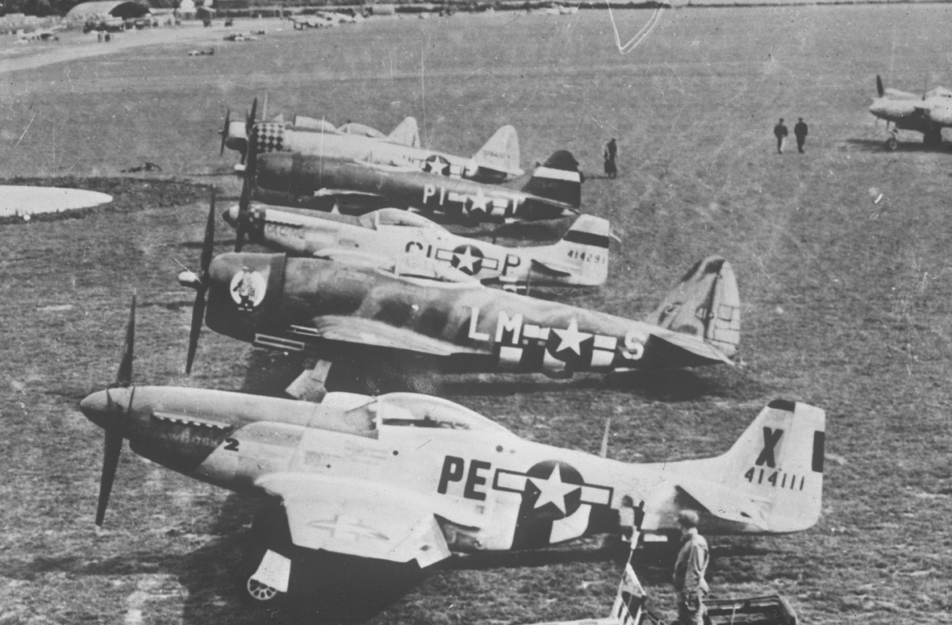 A 328th Fighter Squadron, 352nd Fighter Group P-51 Mustang (PE-X, serial number 44-14111) is at the front of a line of fighter aircraft of various Groups. Behind PE-X is a P-47 Thunderbolt (LM-S) of the 62nd Fighter Squadron, 56th Fighter Group; behind that is a P-51 Mustang (CL-P, serial number 44-14291) of the 338th Fighter Squadron, 55th Fighter Group; and behind that is a P-47 Thunderbolt (PI-T) of the 360th Fighter Squadron, 356th Fighter Group. The line of fighter aircraft continues beyond. Handwrit