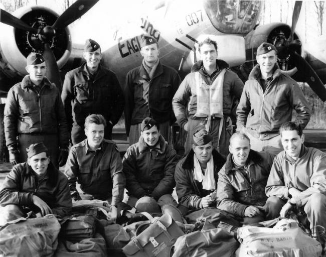 Eleven men in Air Force uniform posed in front of a military airplane.  Five men standing to the rear, six men kneeling in front.  Large canvas bags are on the ground in front of kneeling men.