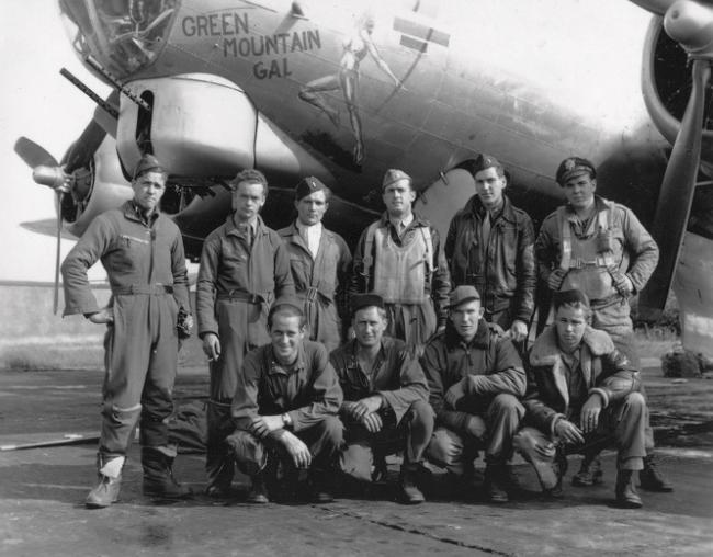 Ten men in Air Force uniform posed in front of a military airplane. Six men standing to the rear, four men kneeling in front.  The men are wearing combat flying gear. 