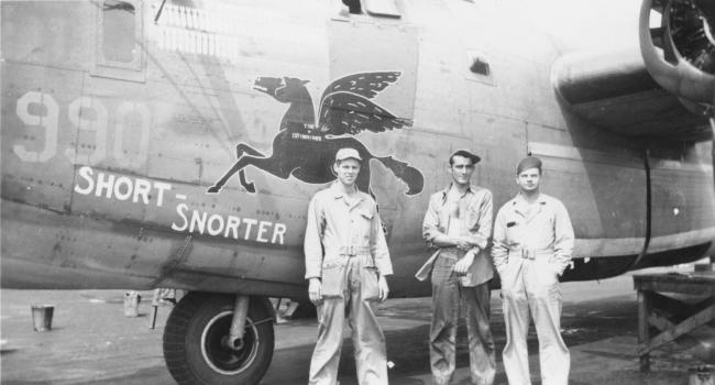 Three airmen of the 392nd Bomb Group with a 597th Bomb Squadron B-24 Liberator (serial number 42-99990) nicknamed "Short Snorter". Jesse Litz is standing in front of the pegasus insignia. Handwritten caption on reverse: '392/597. Jesse LITZ under horse insignia, other two unknown.'