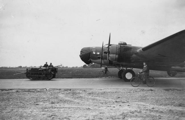 A Cletrac tows a B-17 Flying Fortress (serial number 41-9042) nicknamed "The Berlin Sleeper" of the 92nd Bomb Group down the runway, whilst cyclists guide the aircraft. Passed for publication 17 Oct 1942. Printed caption on reverse: 'New "Fortress" Makes History In The Air. Oct. 1942. The most striking development of the war in the air is undoubtedly the sensational success of the American "Boeing" Flying Fortresses in the daylight bombing of enemy-occupied territory. In the most recent operation 105 Germ